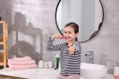 Photo of Cute girl brushing her teeth and showing thumbs up on countertop in bathroom, selective focus