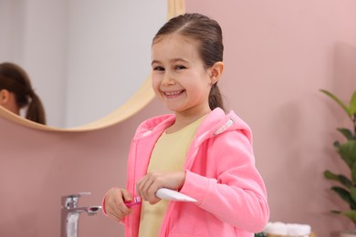 Photo of Cute girl applying toothpaste onto toothbrush in bathroom