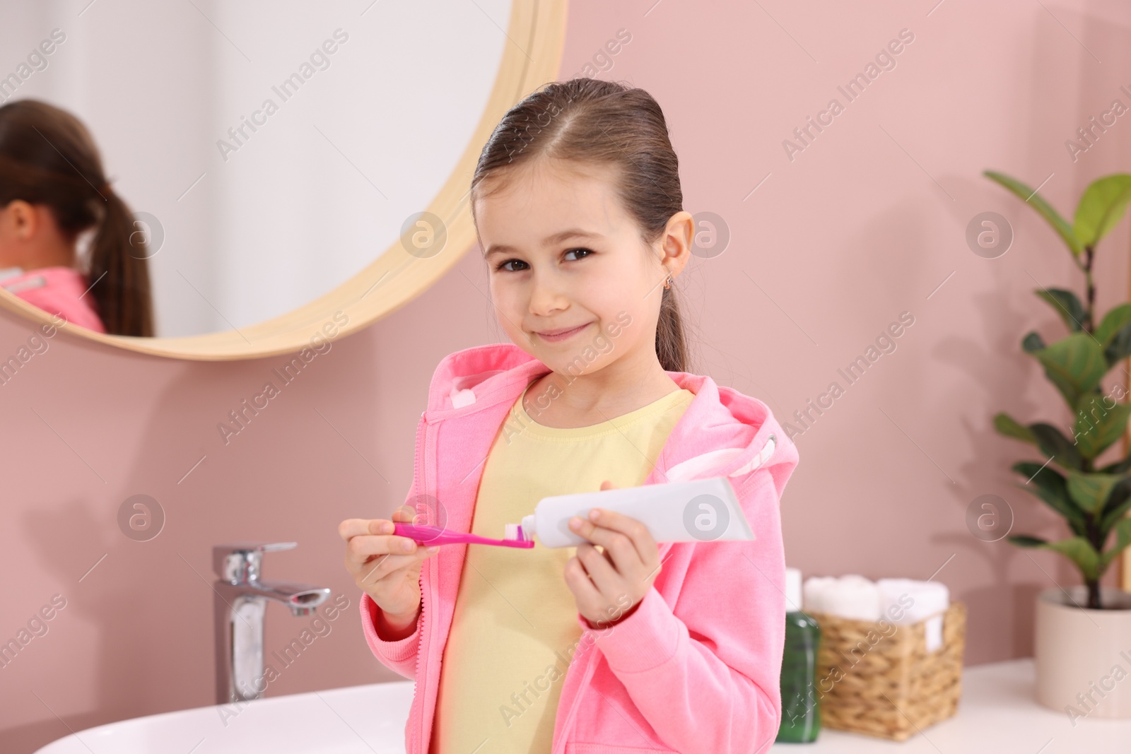 Photo of Cute girl applying toothpaste onto toothbrush in bathroom