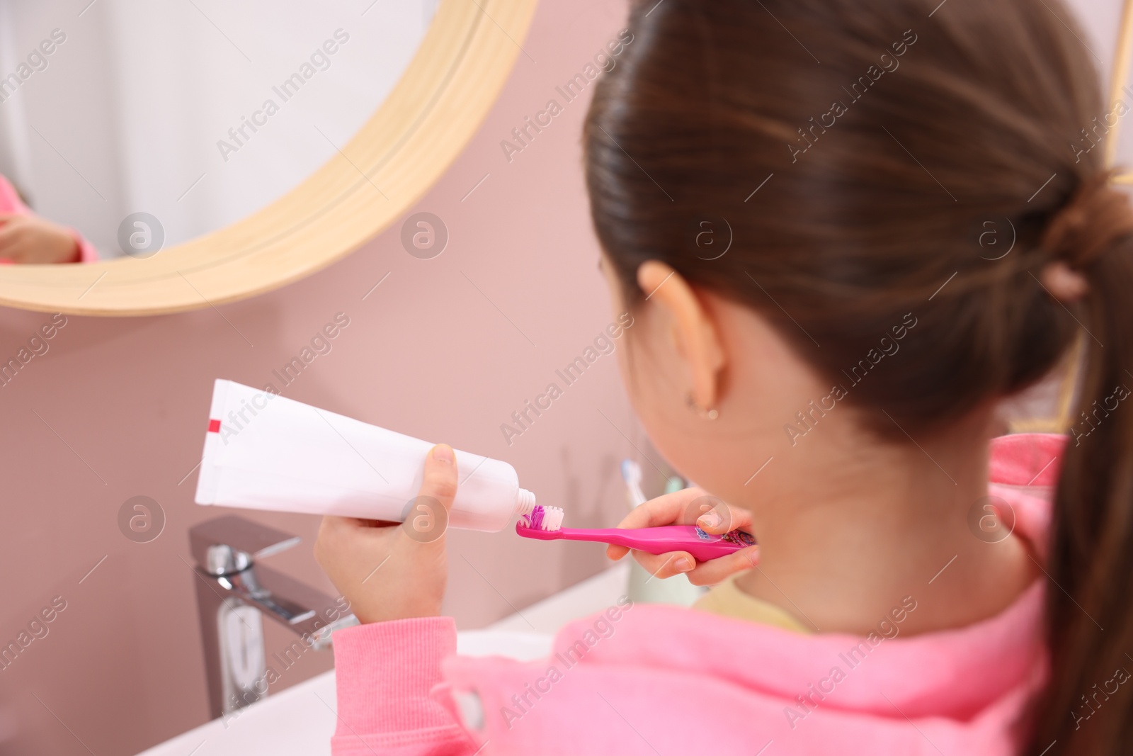 Photo of Cute girl applying toothpaste onto toothbrush in bathroom, back view