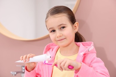 Photo of Cute girl applying toothpaste onto toothbrush in bathroom