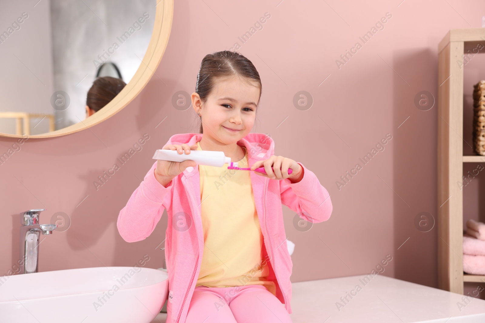 Photo of Cute girl applying toothpaste onto toothbrush in bathroom