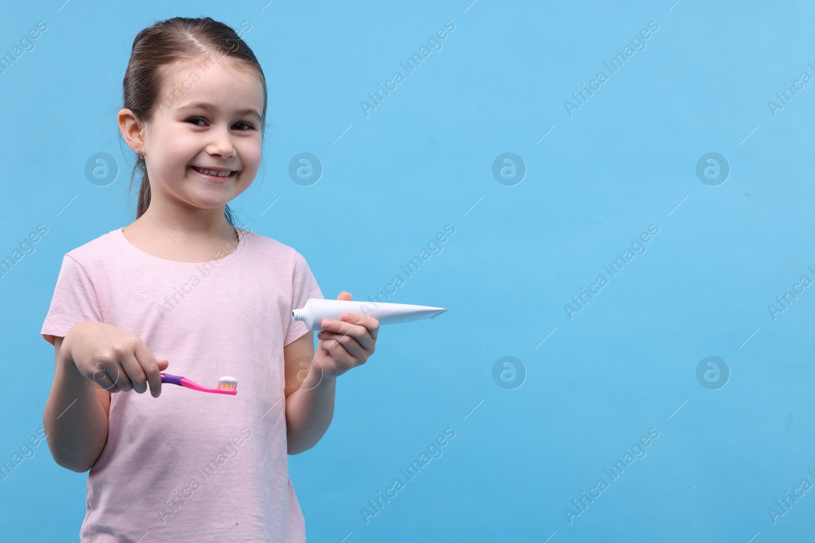 Photo of Cute girl with toothbrush and tube of toothpaste on light blue background, space for text