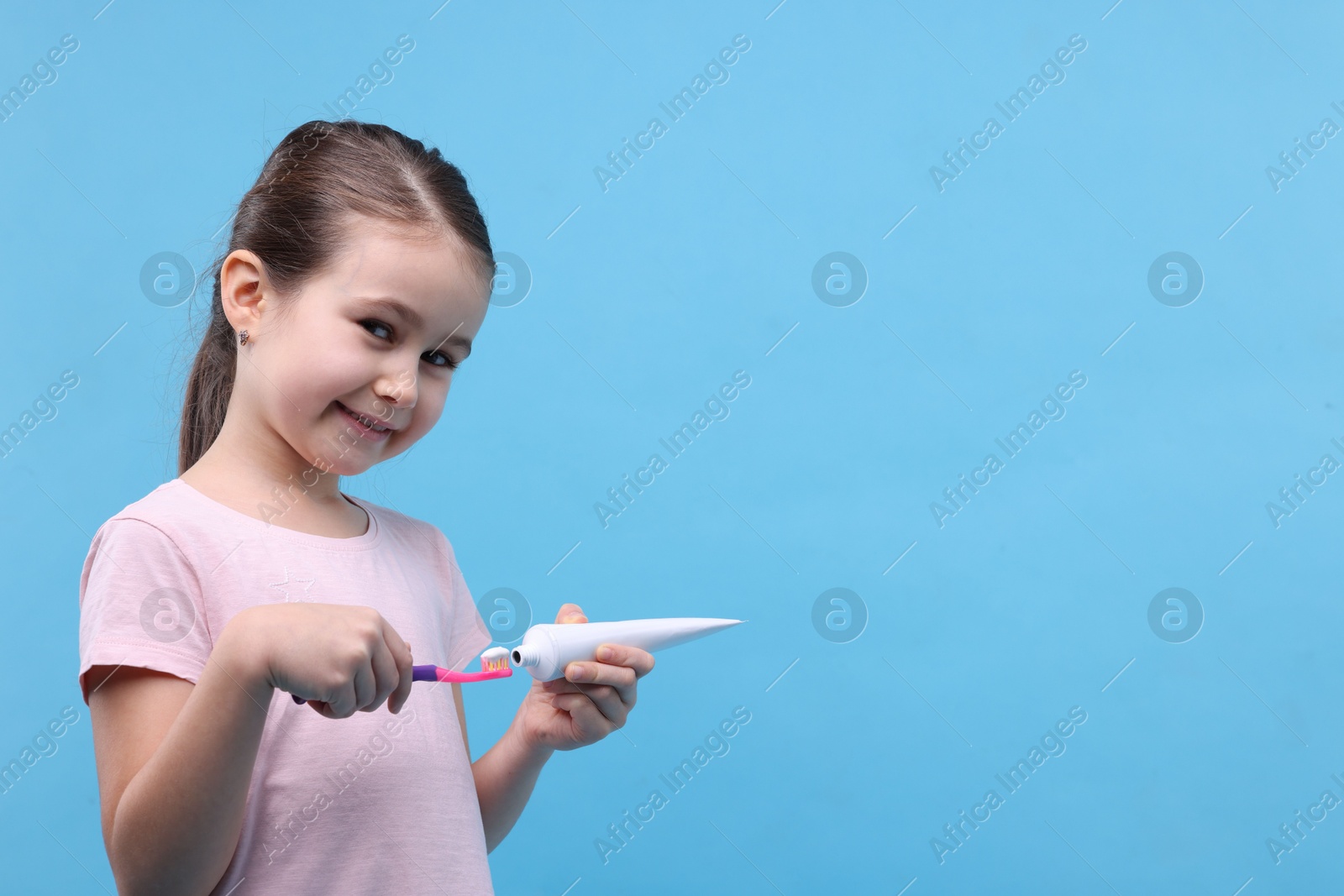 Photo of Cute girl applying toothpaste onto toothbrush on light blue background, space for text