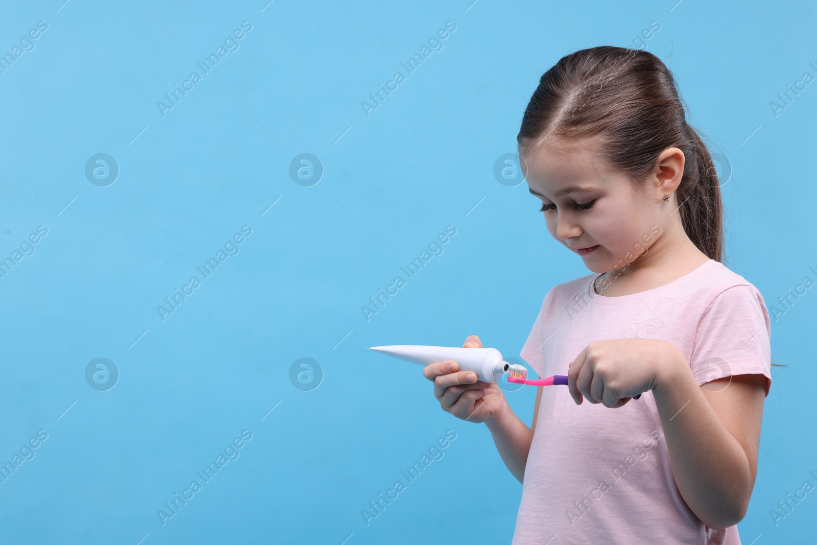 Photo of Cute girl applying toothpaste onto toothbrush on light blue background, space for text