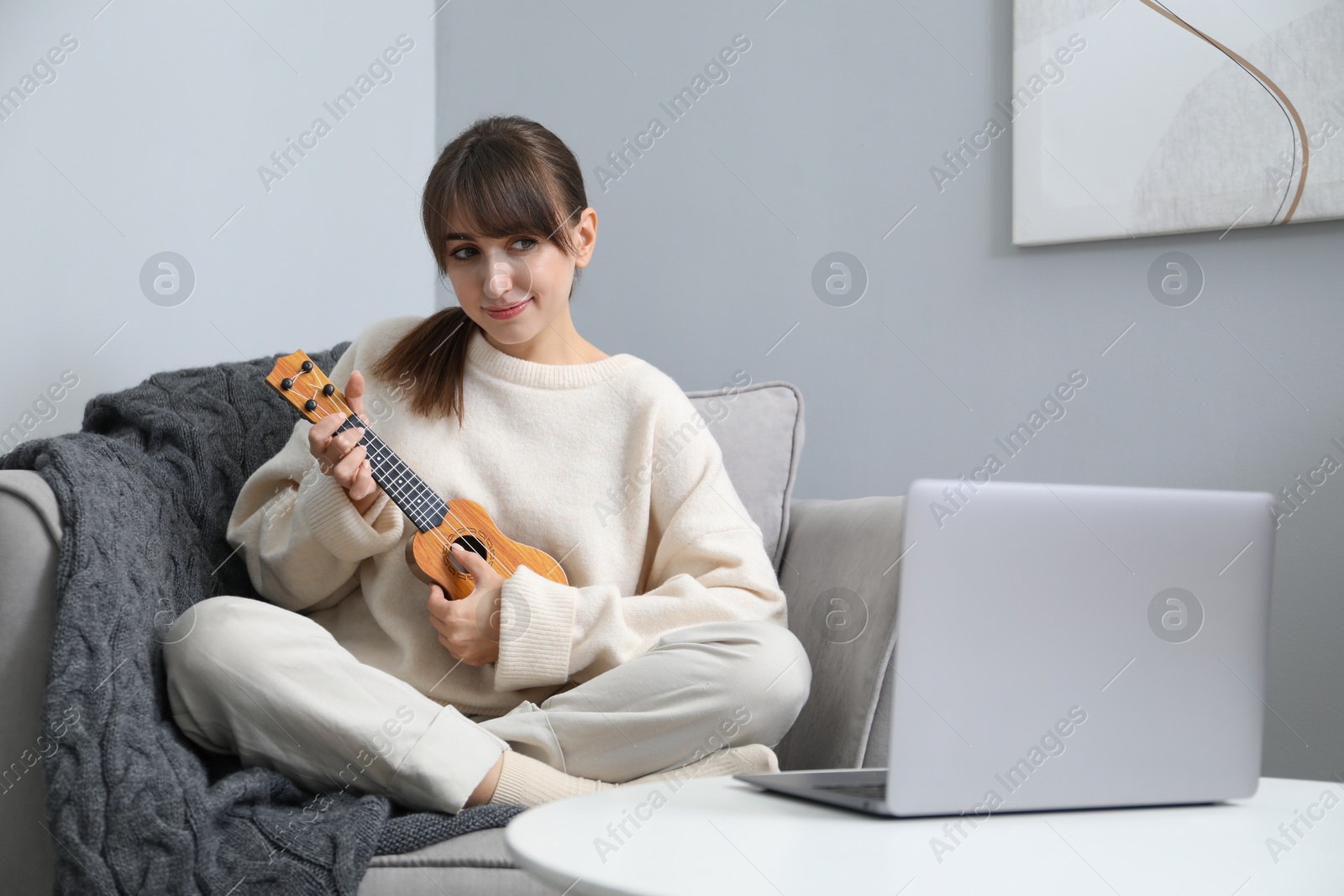 Photo of Woman learning to play ukulele with online music course in armchair at home