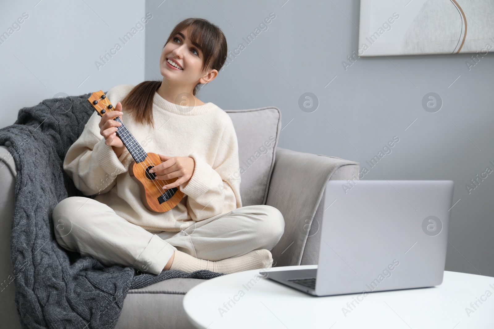 Photo of Woman learning to play ukulele with online music course in armchair at home