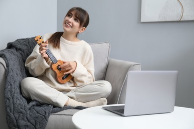 Photo of Woman learning to play ukulele with online music course in armchair at home