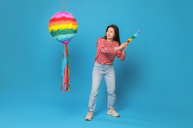 Happy woman hitting colorful pinata with stick on light blue background