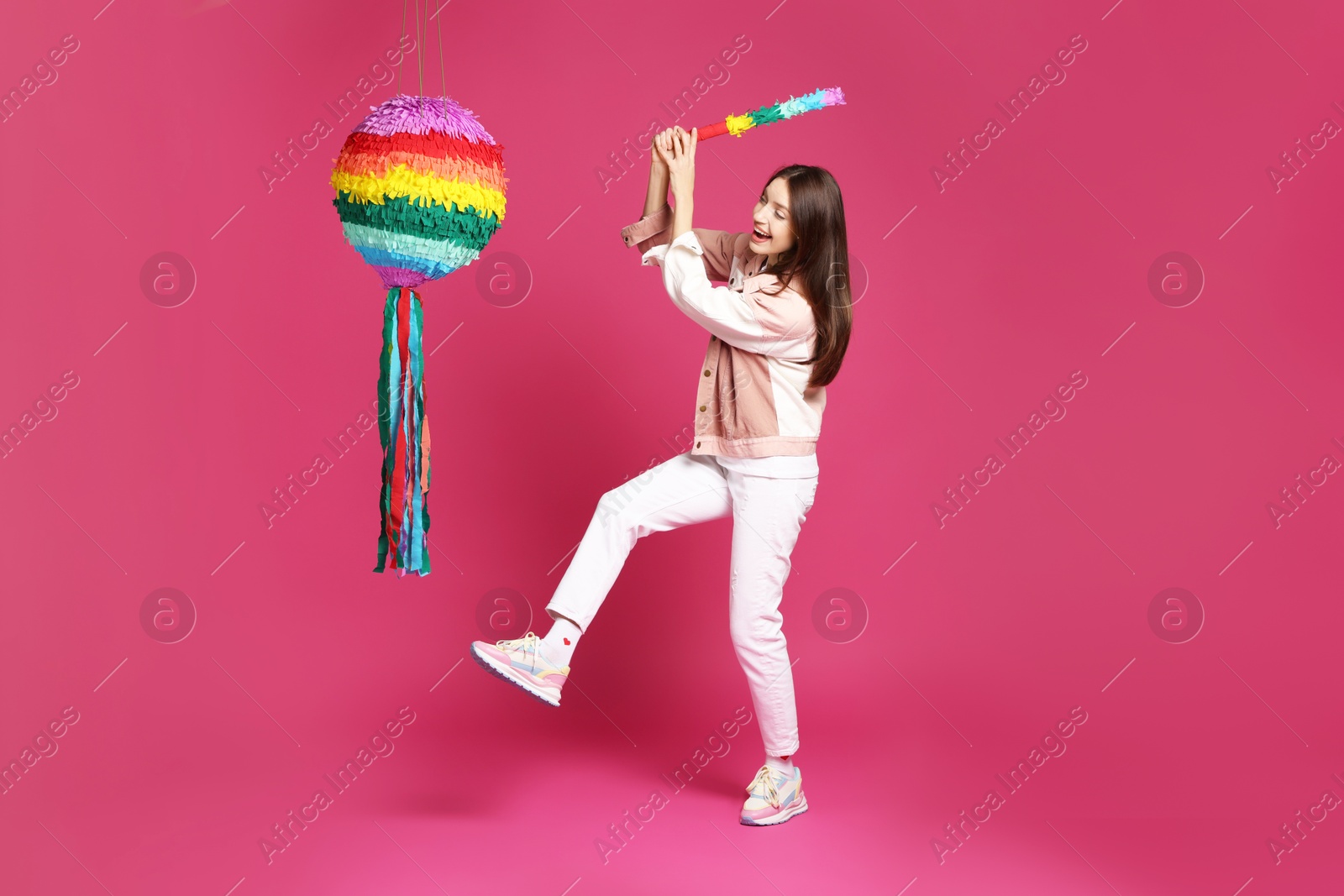 Photo of Happy woman breaking colorful pinata with stick on pink background