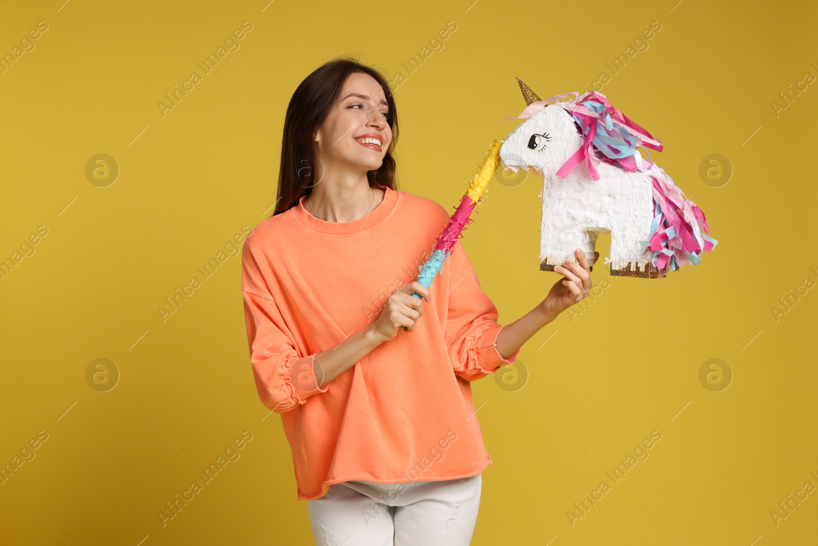 Photo of Happy woman with unicorn shaped pinata and stick on yellow background