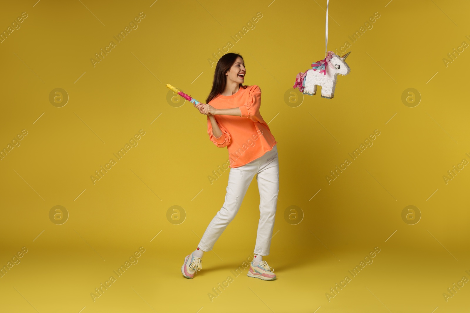 Photo of Happy woman breaking unicorn shaped pinata with stick on yellow background