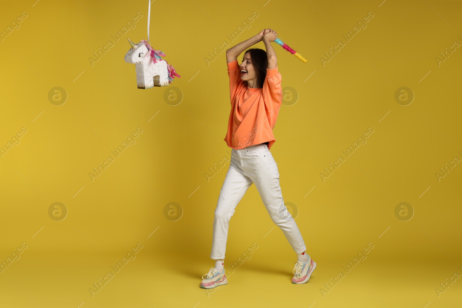 Photo of Happy woman breaking unicorn shaped pinata with stick on yellow background