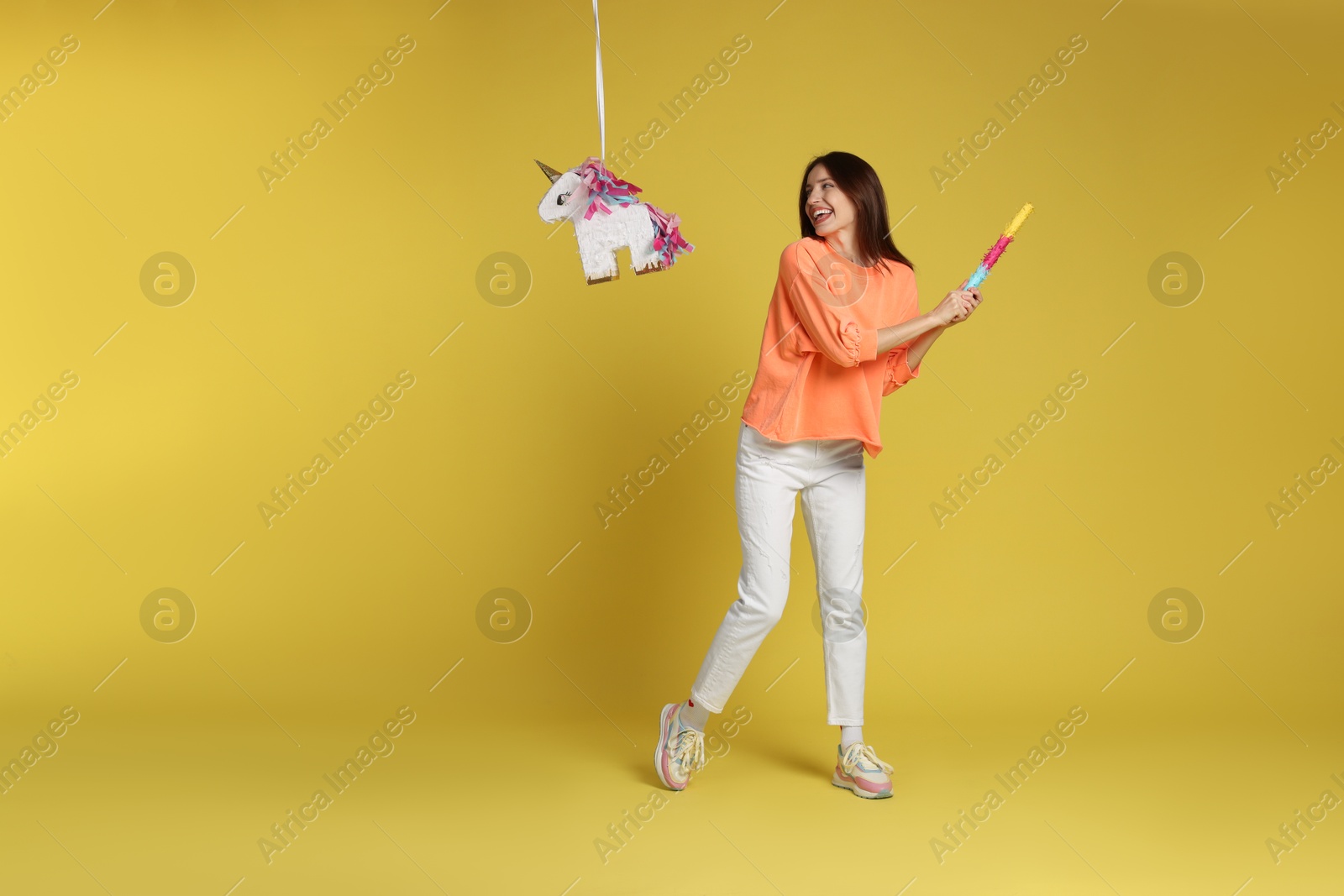 Photo of Happy woman breaking unicorn shaped pinata with stick on yellow background