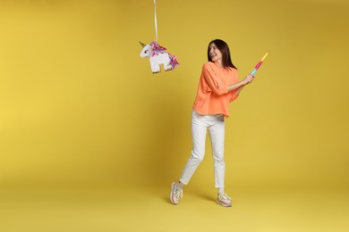 Happy woman breaking unicorn shaped pinata with stick on yellow background