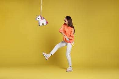 Photo of Happy woman breaking unicorn shaped pinata with stick on yellow background