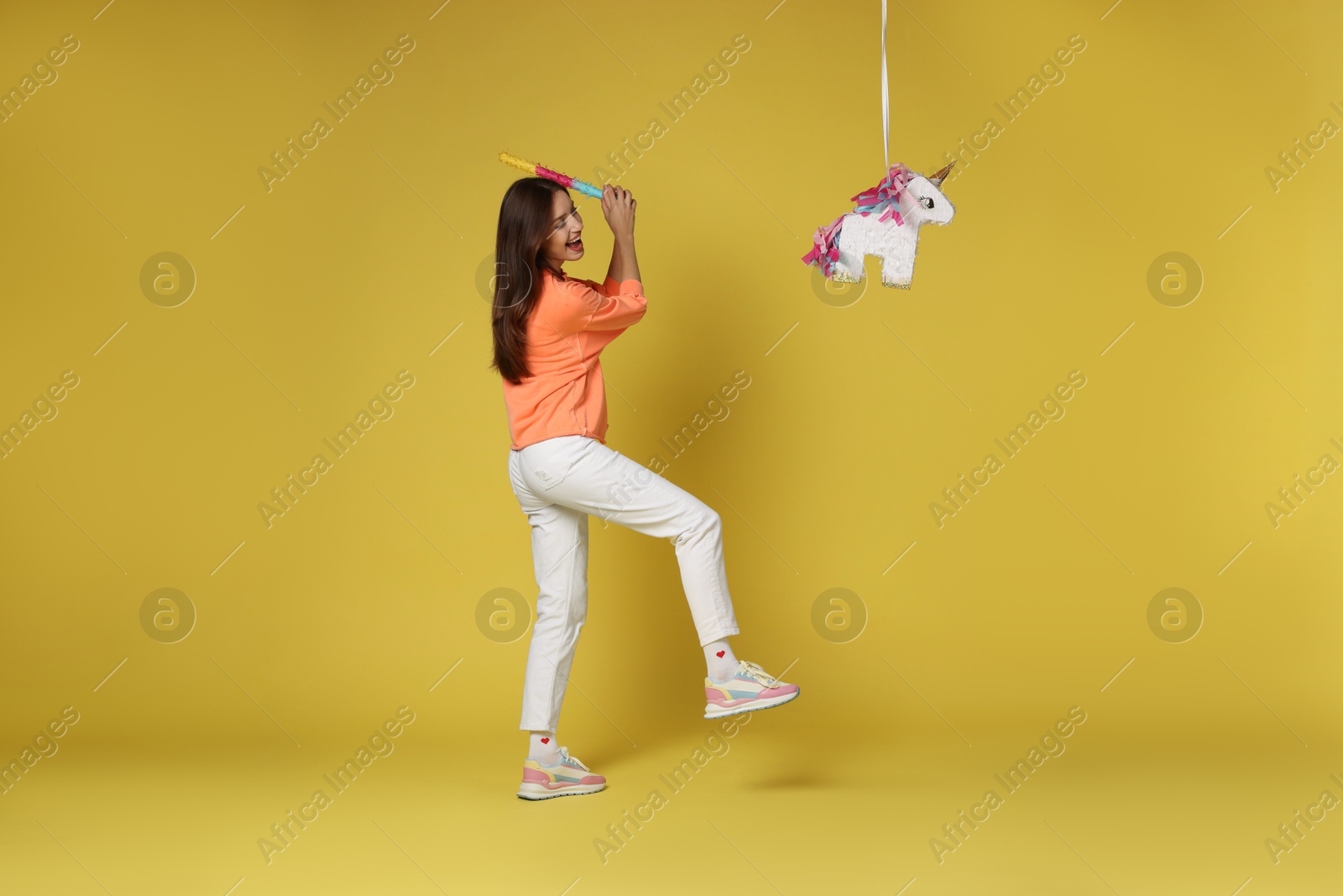 Photo of Happy woman breaking unicorn shaped pinata with stick on yellow background