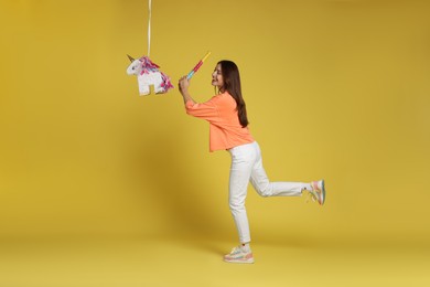 Photo of Happy woman breaking unicorn shaped pinata with stick on yellow background