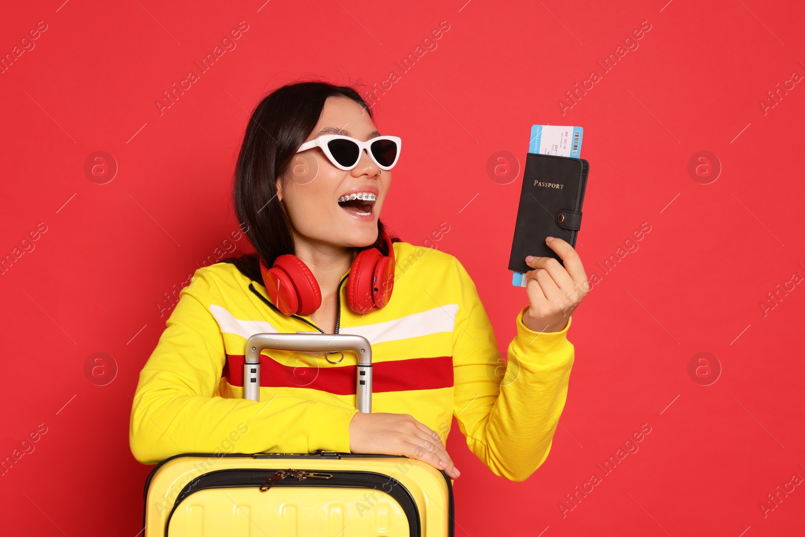 Photo of Happy traveller with headphones, passport and suitcase on red background