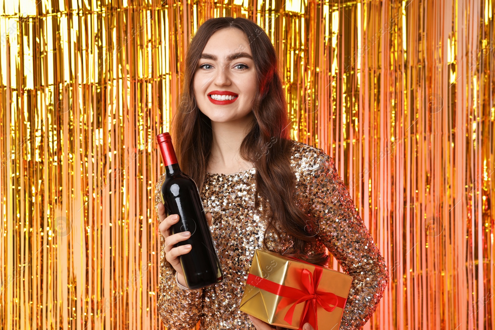 Photo of Happy young woman with gift box and bottle of wine against foil curtain