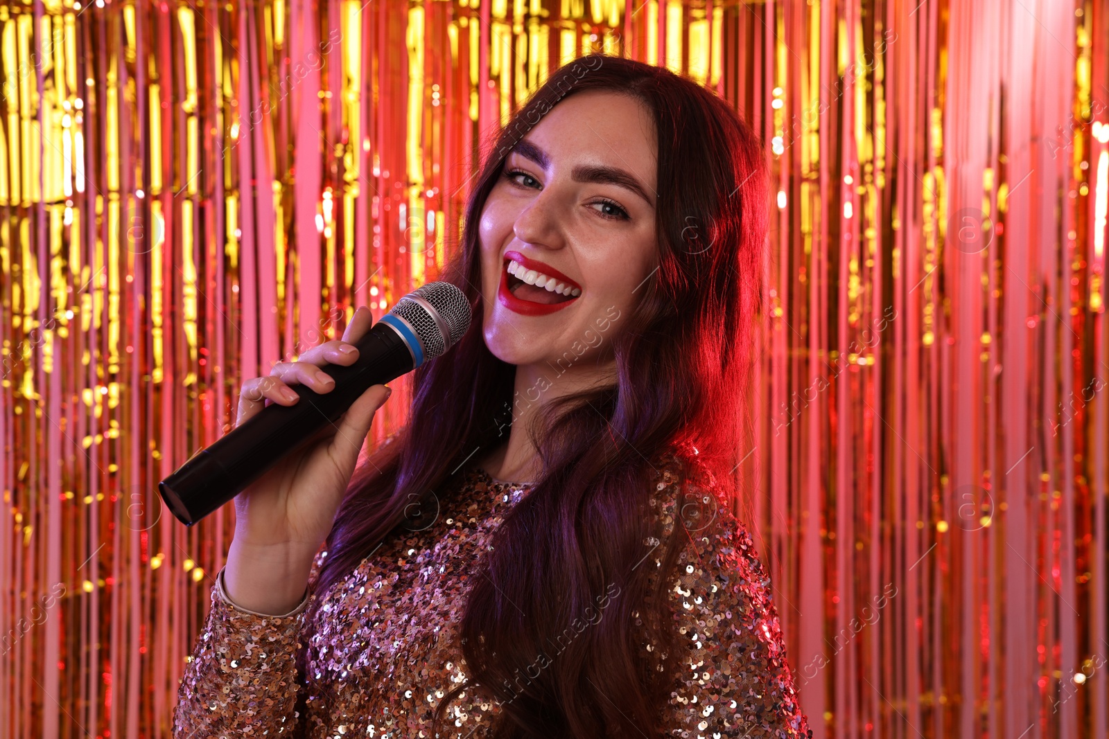 Photo of Young woman with microphone singing against foil curtain in neon lights