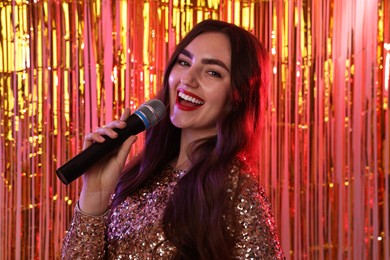 Photo of Young woman with microphone singing against foil curtain in neon lights