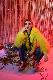 Photo of Beautiful young woman wearing retro outfit with tinsel and disco balls against golden foil curtain in neon lights