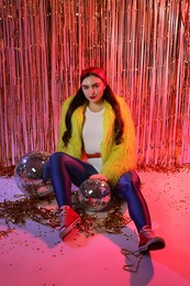 Photo of Beautiful young woman wearing retro outfit with tinsel and disco balls against golden foil curtain in neon lights