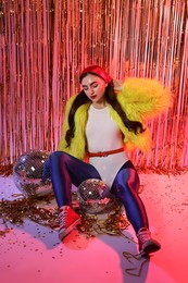 Photo of Beautiful young woman wearing retro outfit with tinsel and disco balls against golden foil curtain in neon lights