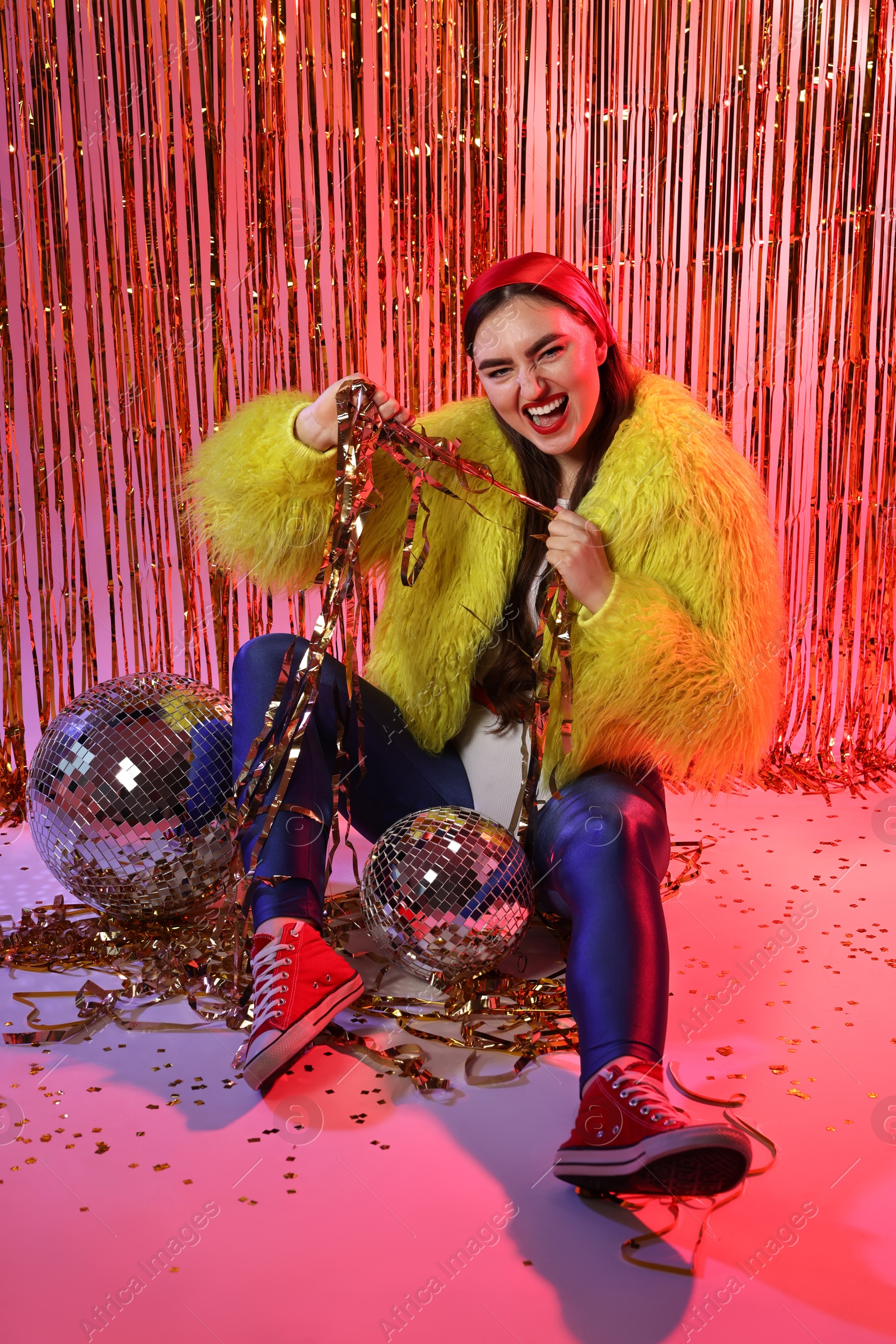 Photo of Beautiful young woman wearing retro outfit with tinsel and disco balls against golden foil curtain in neon lights