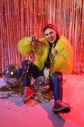 Photo of Beautiful young woman wearing retro outfit with tinsel and disco balls against golden foil curtain in neon lights