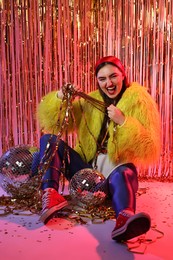 Photo of Beautiful young woman wearing retro outfit with tinsel and disco balls against golden foil curtain in neon lights