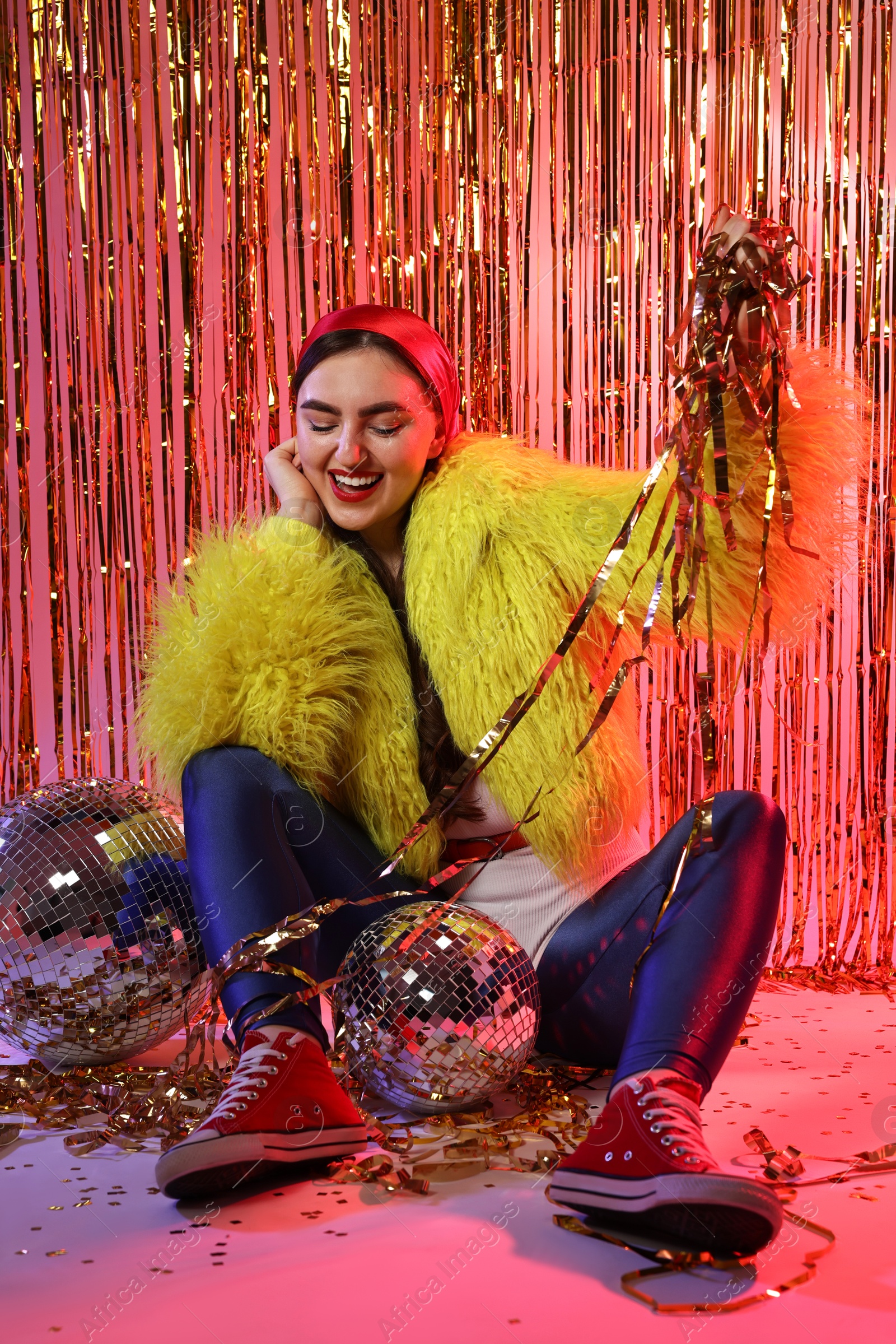 Photo of Beautiful young woman wearing retro outfit with tinsel and disco balls against golden foil curtain in neon lights