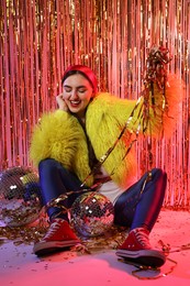 Photo of Beautiful young woman wearing retro outfit with tinsel and disco balls against golden foil curtain in neon lights