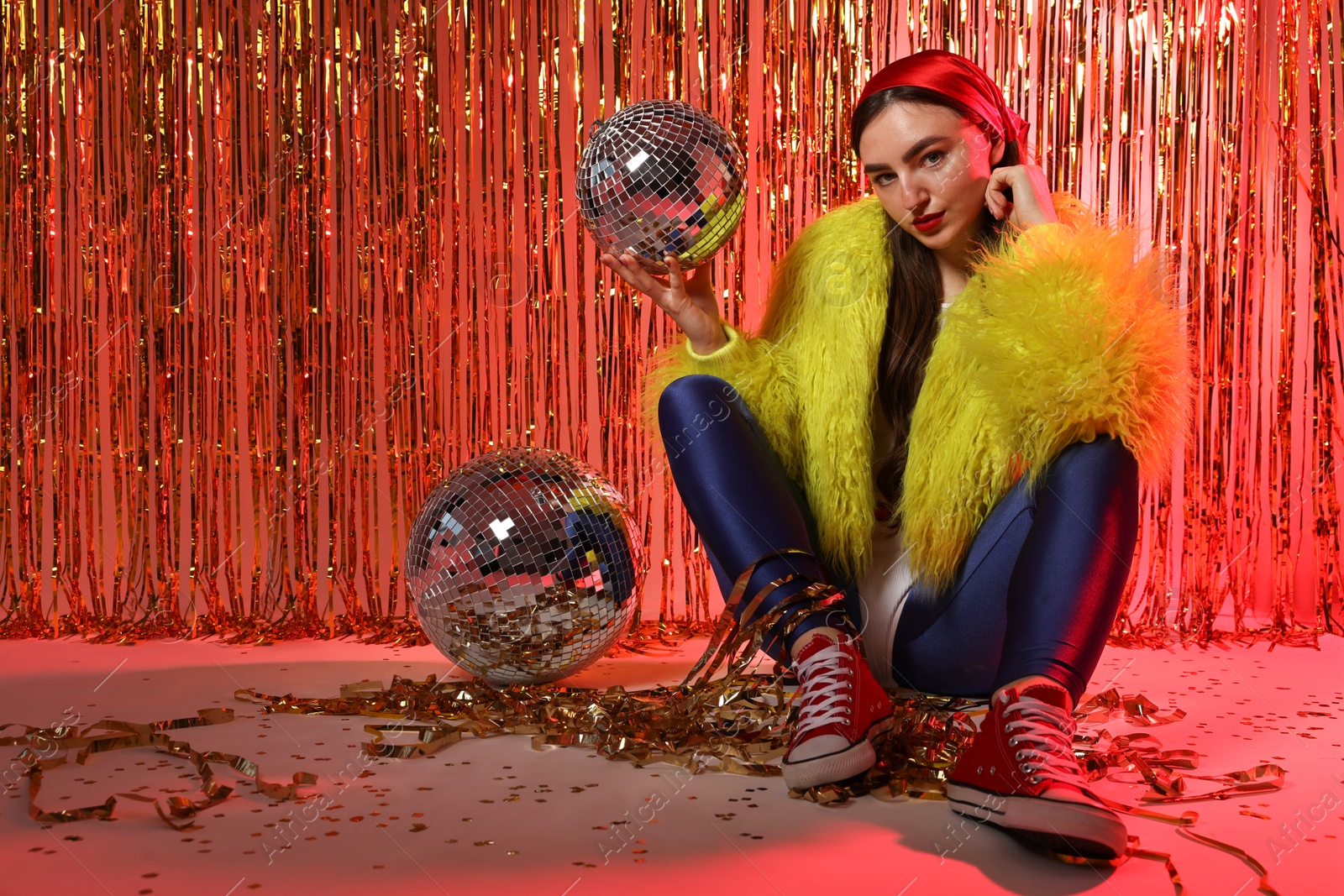 Photo of Beautiful young woman wearing retro outfit with tinsel and disco balls against golden foil curtain in neon lights