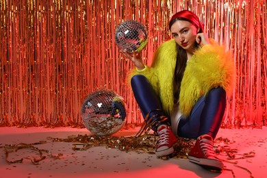 Photo of Beautiful young woman wearing retro outfit with tinsel and disco balls against golden foil curtain in neon lights