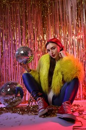 Photo of Beautiful young woman wearing retro outfit with tinsel and disco balls against golden foil curtain in neon lights