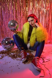 Photo of Beautiful young woman wearing retro outfit with tinsel and disco balls against golden foil curtain in neon lights