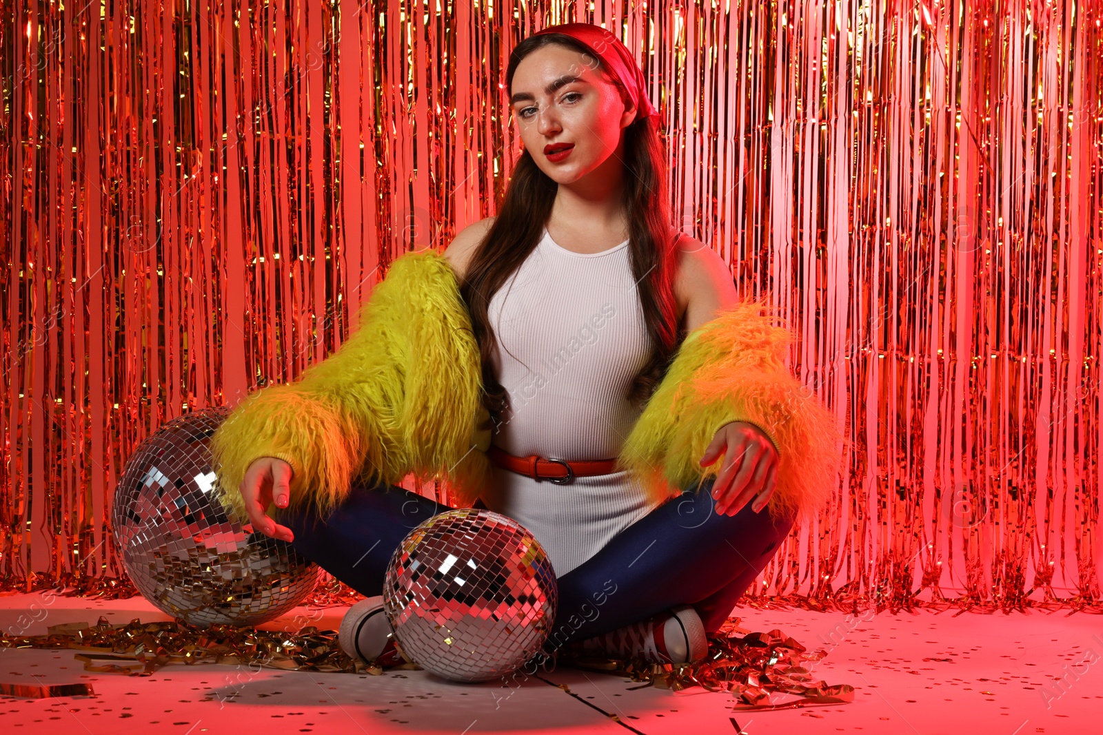 Photo of Beautiful young woman wearing retro outfit with tinsel and disco balls against golden foil curtain in neon lights