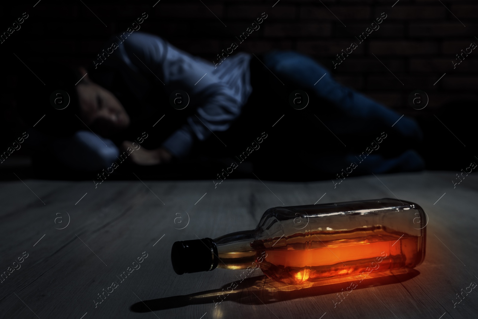 Image of Alcohol addiction. Drunk man lying on floor, focus on bottle of drink in darkness