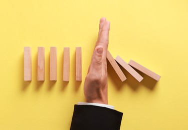 Photo of Man stopping wooden blocks from falling on yellow background, top view. Domino effect