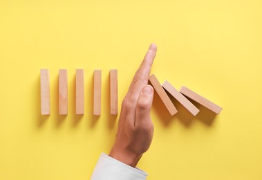 Photo of Man stopping wooden blocks from falling on yellow background, top view. Domino effect