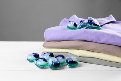 Photo of Laundry detergent capsules and clean clothes on white wooden table, closeup. Space for text