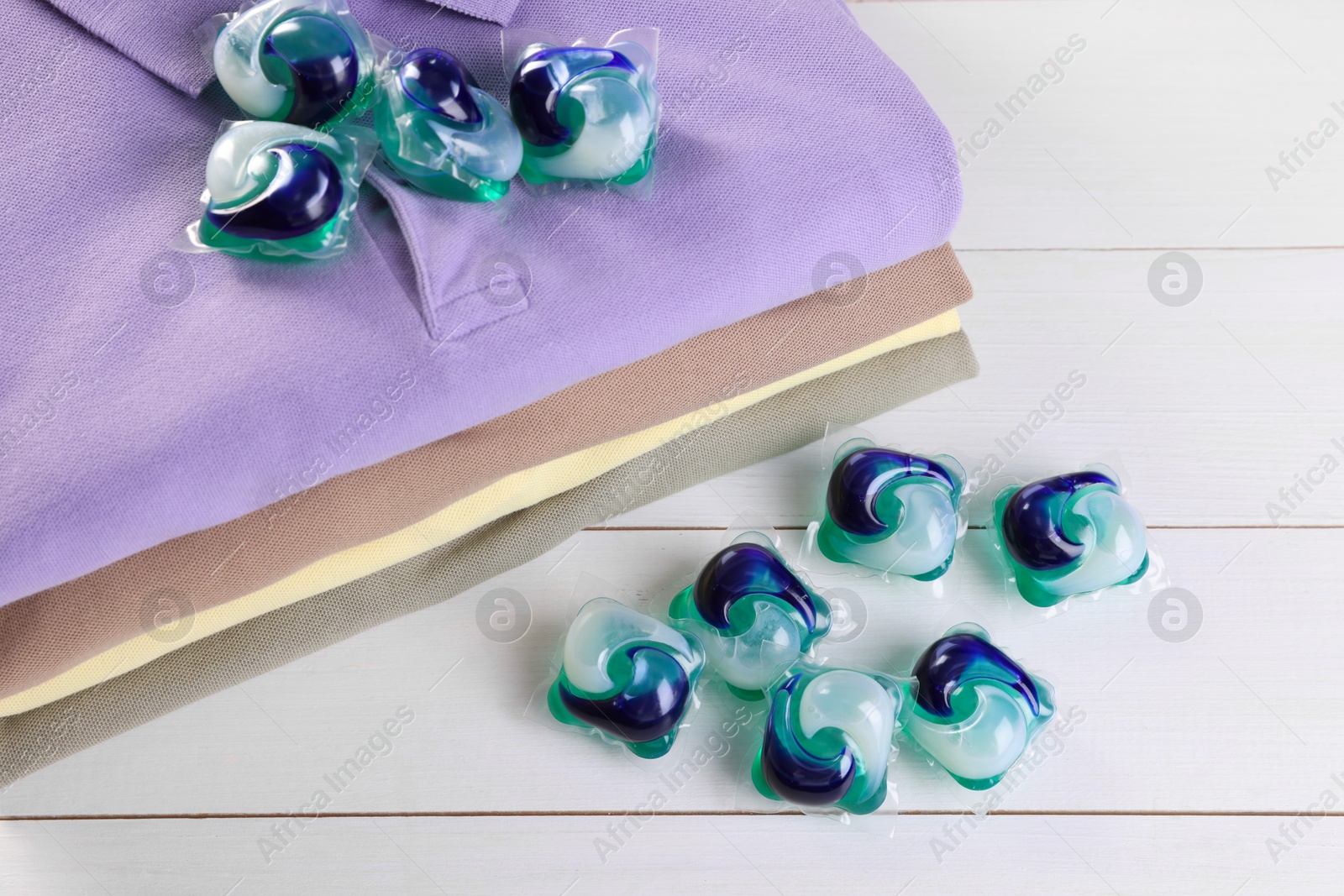 Photo of Laundry detergent capsules and clean clothes on white wooden table, closeup