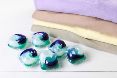 Photo of Laundry detergent capsules and clean clothes on white wooden table, closeup