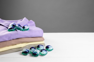 Photo of Laundry detergent capsules and clean clothes on white wooden table. Space for text