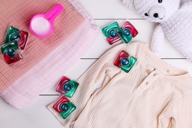 Photo of Laundry detergent capsules, clean clothes and teddy bear on white wooden background, flat lay