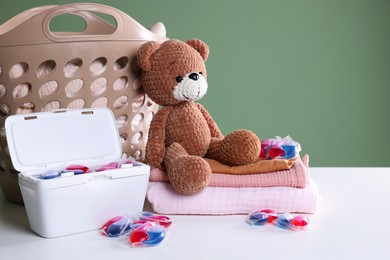 Photo of Detergent capsules, clean linens, teddy bear and laundry basket on white table. Space for text