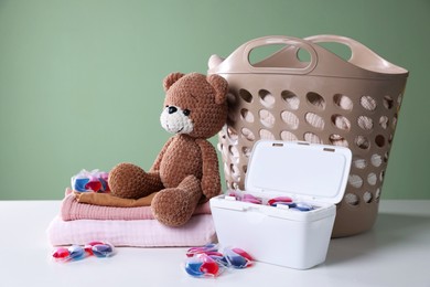 Photo of Detergent capsules, clean linens, teddy bear and laundry basket on white table