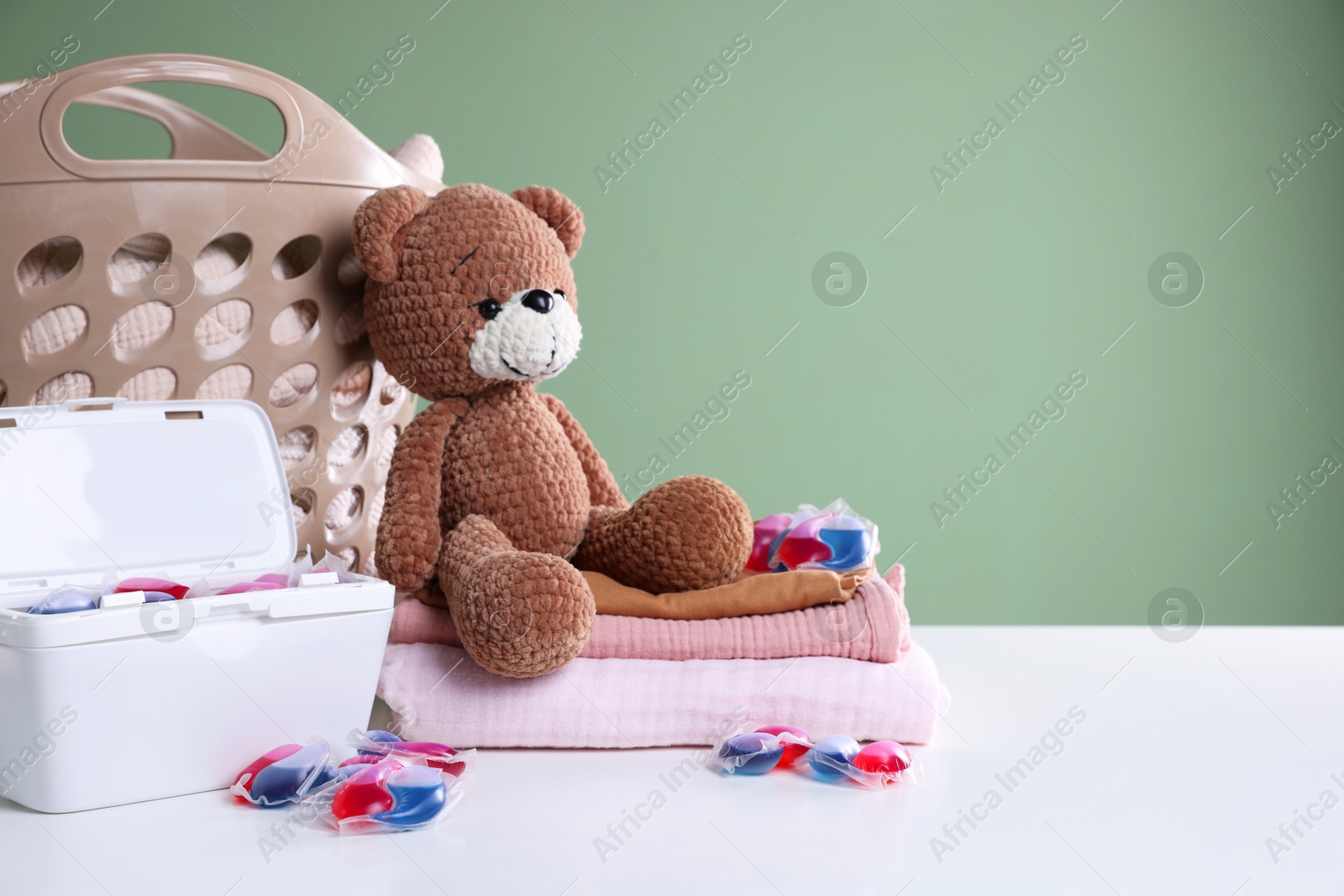 Photo of Detergent capsules, clean linens, teddy bear and laundry basket on white table. Space for text